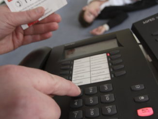 Im Vordergrund sind ein Telefon und eine Hand zu sehen. Im Hintergrund liegt eine Person bewusstlos auf dem Fußboden.