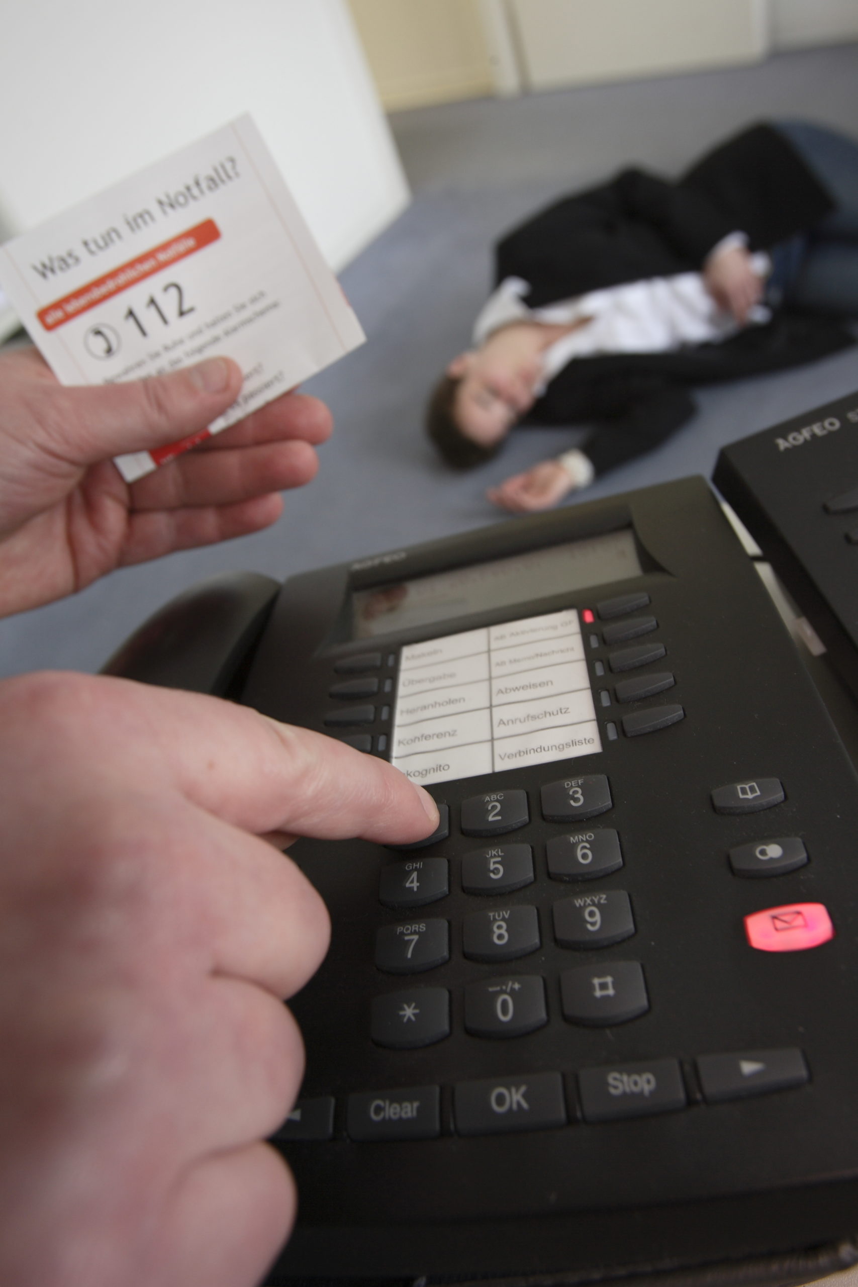 Im Vordergrund sind ein Telefon und eine Hand zu sehen. Im Hintergrund liegt eine Person bewusstlos auf dem Fußboden.