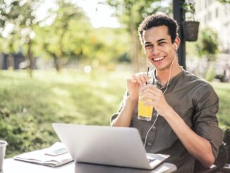 Ein junger Mann sitzt mit einem Drink in der Hand und Kopfhörern im Ohr in der Natur vor einem Laptop.