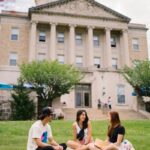 Drei junge Menschen sitzen auf der Wiede vor einem klassizistischen Gebäude