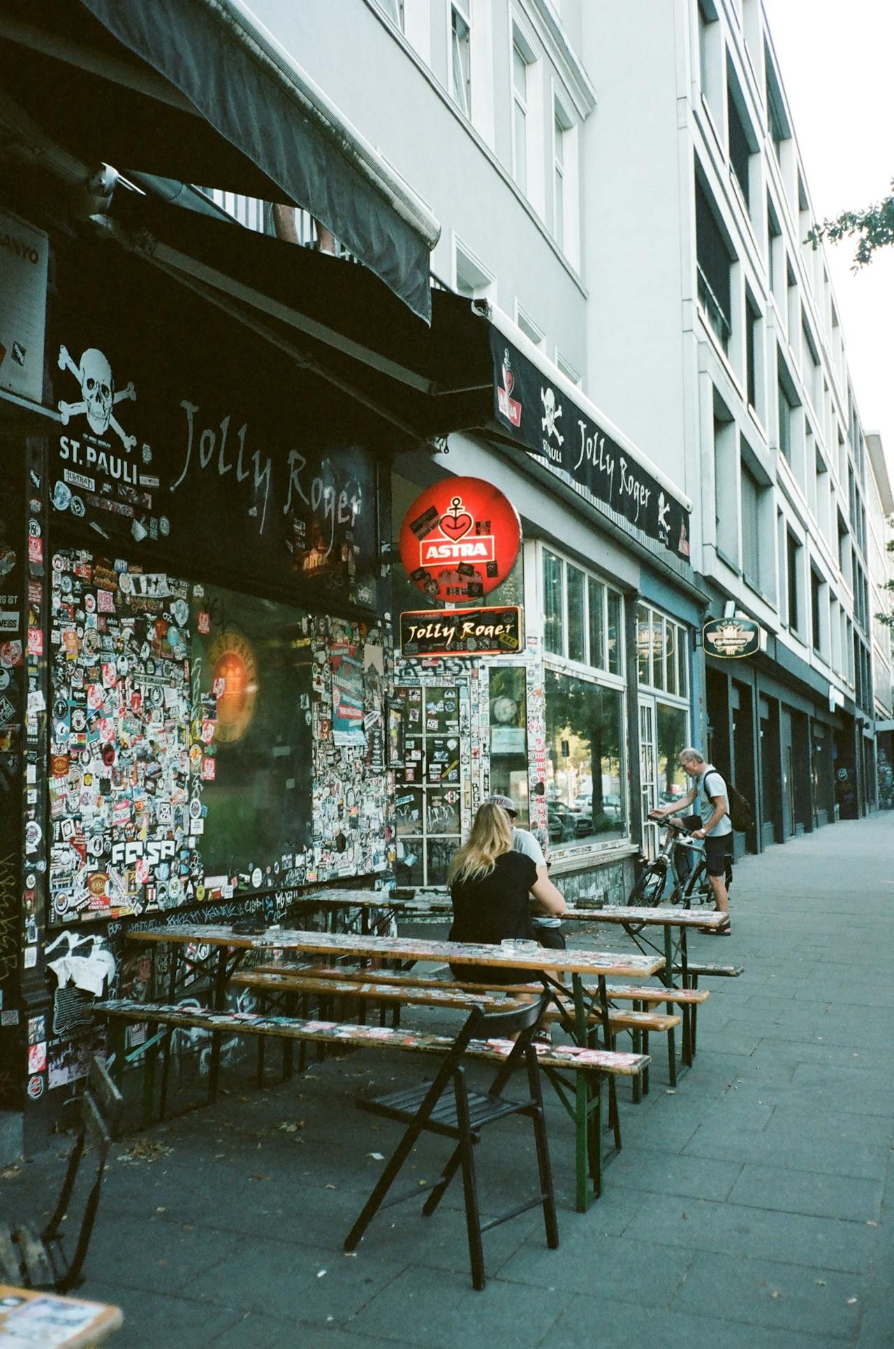 Hamburg St. Pauli: Außenansicht der Bar Jolly Roger mit Gästen an Holzbänken und beklebter Fassade im Tageslicht.