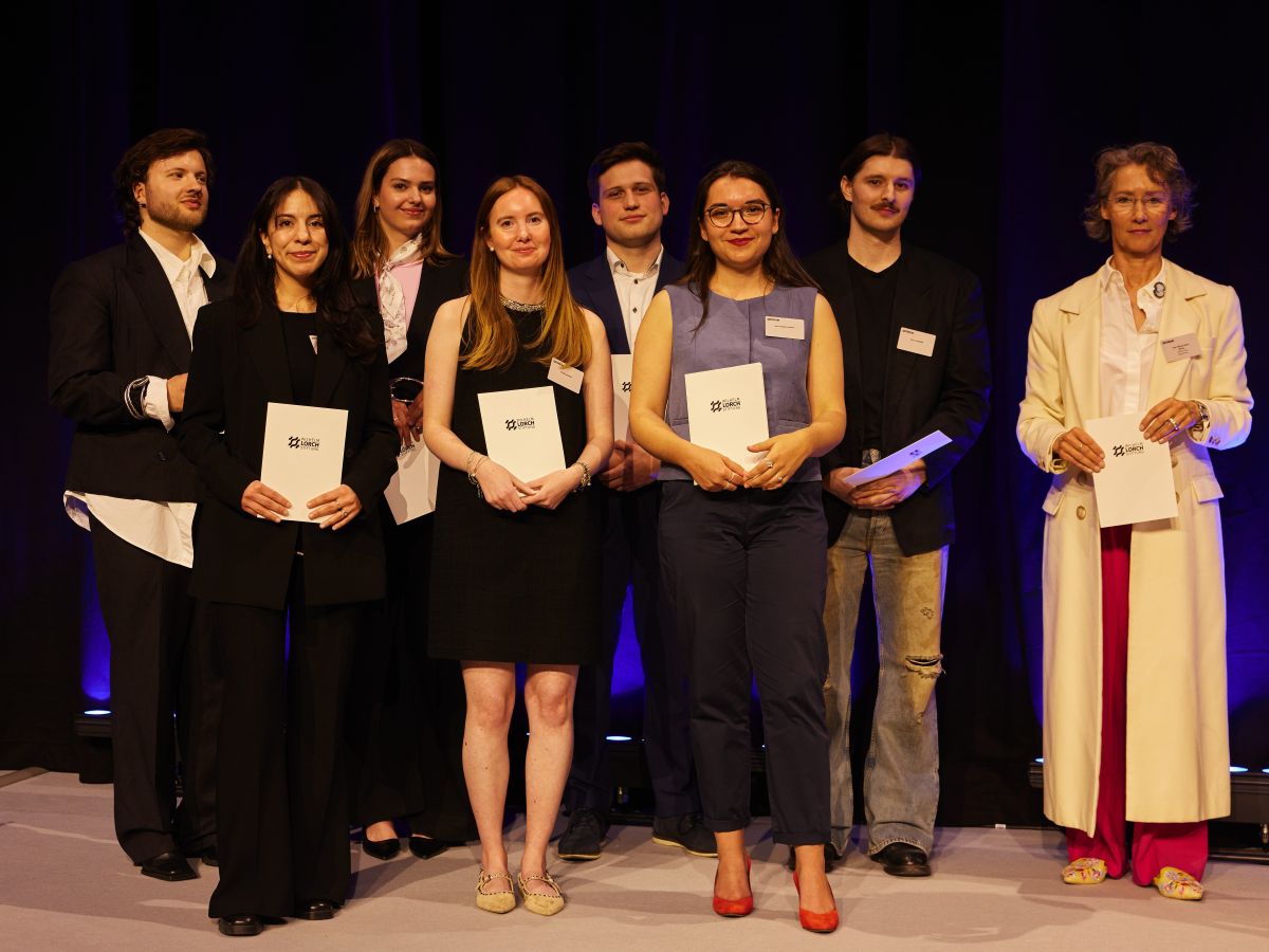 Group of eight young adults on a dimly lit stage, each holding a white certificate or folder, posing for a formal award ceremony photograph.
