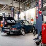 Blue vintage car on a workshop floor with open rear hatch, surrounded by car lifts and equipment in a garage.