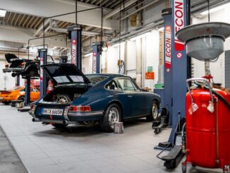 Blue vintage car on a workshop floor with open rear hatch, surrounded by car lifts and equipment in a garage.