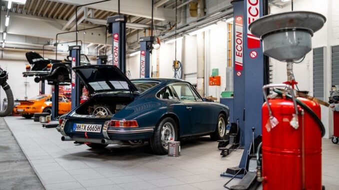 Blue vintage car on a workshop floor with open rear hatch, surrounded by car lifts and equipment in a garage.