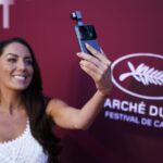 Woman at a film festival takes a selfie with a phone mounted to a grip, in front of a Cannes backdrop.