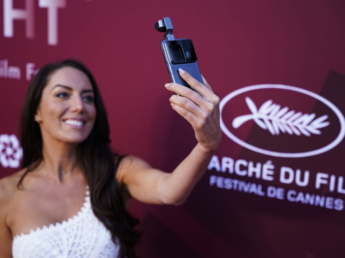 Woman at a film festival takes a selfie with a phone mounted to a grip, in front of a Cannes backdrop.