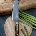 Chef's knife with a wooden handle rests on a round cutting board beside fresh asparagus spears.