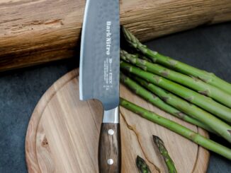 Chef's knife with a wooden handle rests on a round cutting board beside fresh asparagus spears.