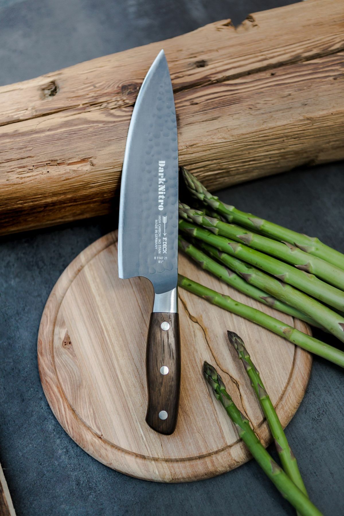 Chef's knife with a wooden handle rests on a round cutting board beside fresh asparagus spears.