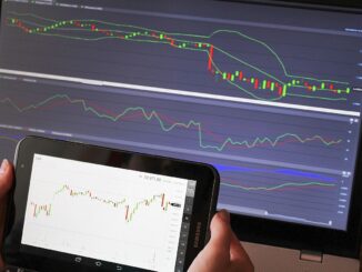 Two screens show stock charts; a tablet in the foreground displays a candlestick chart (DAX) while a laptop monitor in the background shows multiple green and red lines.