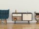Blue tufted chair with wooden legs sits next to a two-door black-framed sideboard on a light wall.