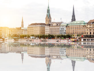 Historic city skyline along a river with ornate towers and pastel rooftops, reflected in calm water, sunset glow nearby.