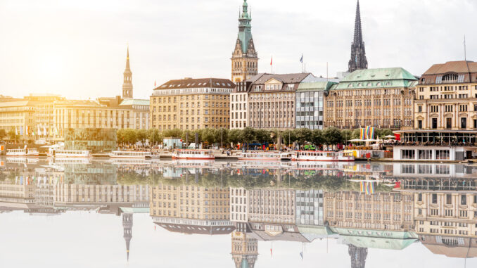 Historic city skyline along a river with ornate towers and pastel rooftops, reflected in calm water, sunset glow nearby.