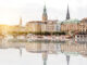 Historic city skyline along a river with ornate towers and pastel rooftops, reflected in calm water, sunset glow nearby.