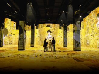 Group of four visitors standing in a gold-toned gallery with large mirrored pillars and Klimt-inspired wall panels ahead.