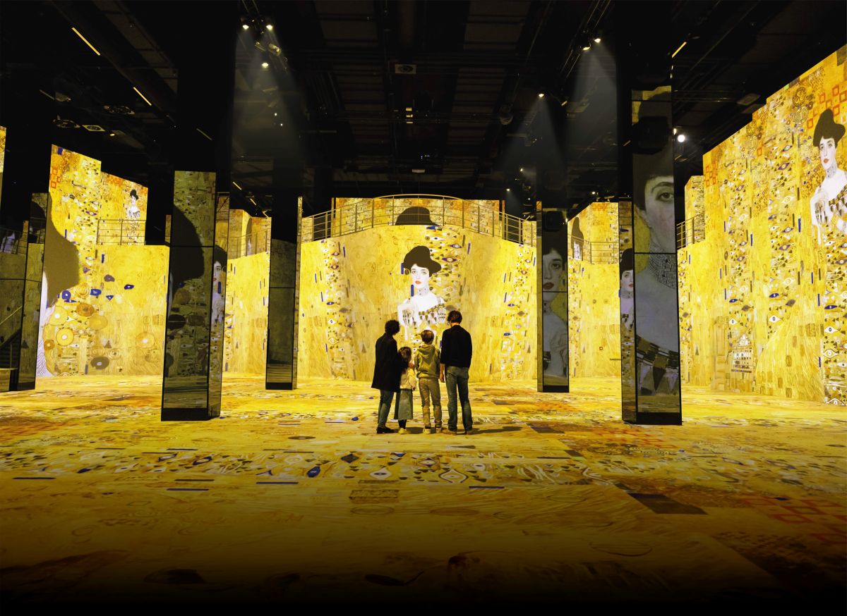 Group of four visitors standing in a gold-toned gallery with large mirrored pillars and Klimt-inspired wall panels ahead.