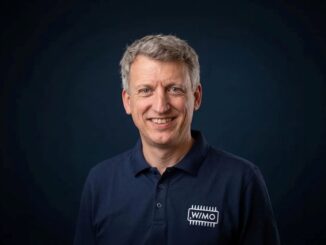 Portrait of a smiling middle-aged man with gray hair wearing a navy polo shirt with a WIMO logo on a dark background.