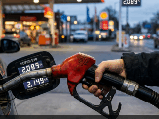 Close-up of a hand gripping a red gas pump nozzle at a fueling station, with a blurred convenience store and cars in the background.