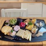 Cheese board on a black tray with assorted cheeses, grapes, and small bowls of cubes on a wooden table.