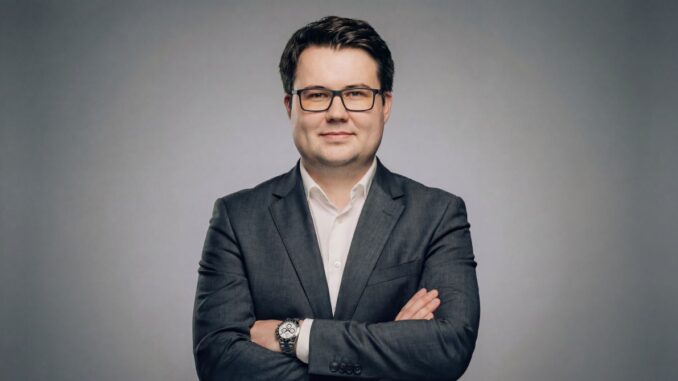 Portrait of a man in a dark suit with a white shirt, arms crossed, wearing glasses, against a gray studio background.