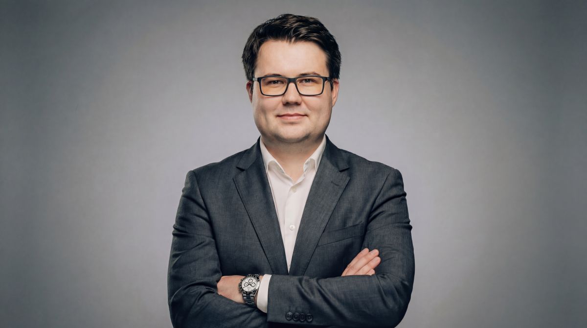 Portrait of a man in a dark suit with a white shirt, arms crossed, wearing glasses, against a gray studio background.