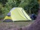 Yellow and gray dome tent pitched on a grassy campsite with trees in the background and camping gear to the right.