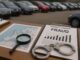 Desk setup for fraud investigation: clipboard labeled FRAUD with a bar chart, handcuffs, magnifying glass, and a map nearby. Vehicles are blurred in the background.