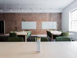 Bright conference room with a brick wall, two long light wood tables, and green upholstered chairs around, large window on the right. In the foreground, a glass cup holding white pencils sits on the table.