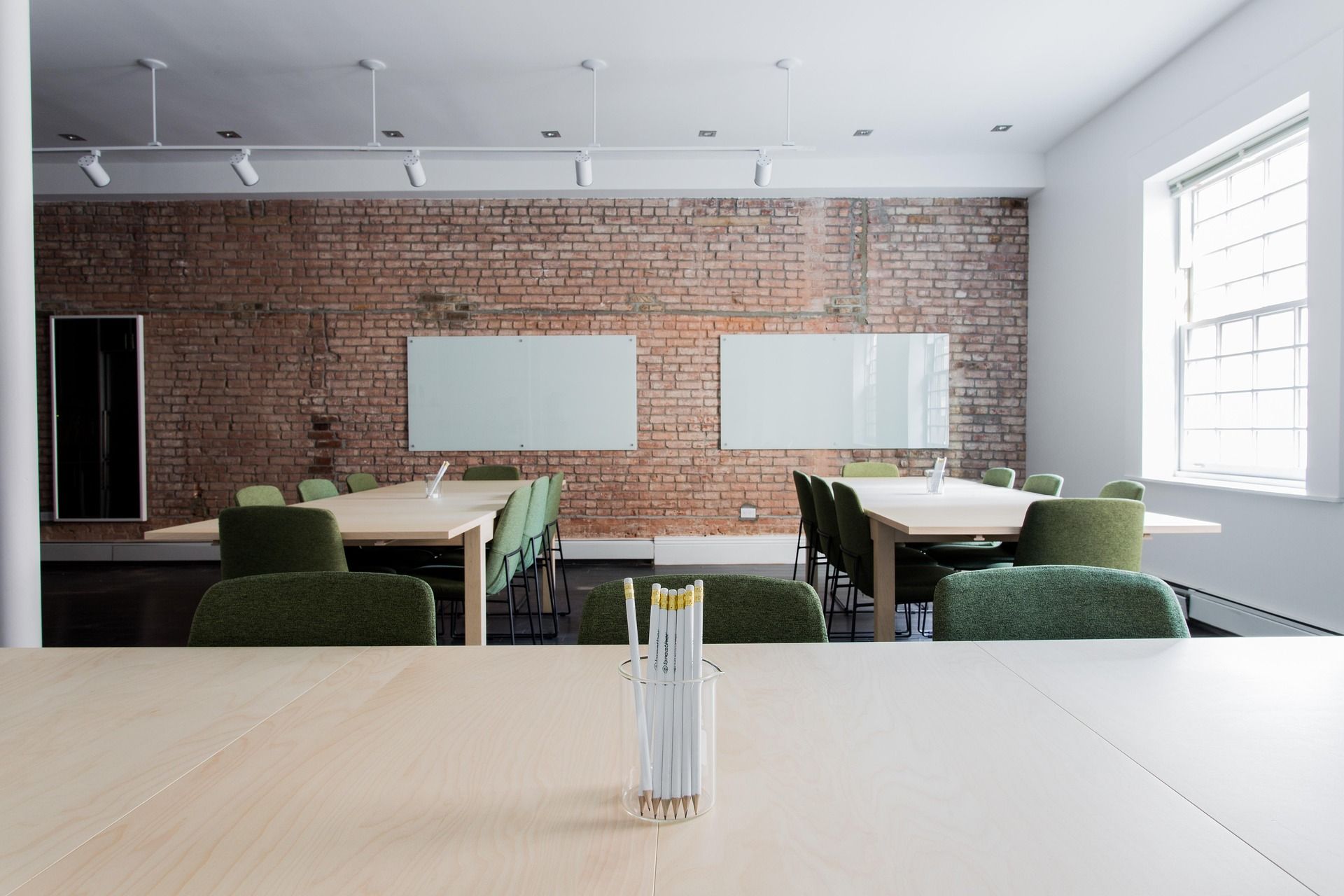 Bright conference room with a brick wall, two long light wood tables, and green upholstered chairs around, large window on the right. In the foreground, a glass cup holding white pencils sits on the table.
