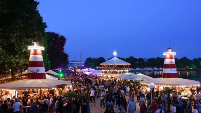 Crowd at an outdoor festival by a waterfront with tents, string lights, and two red-and-white lighthouse structures at dusk.