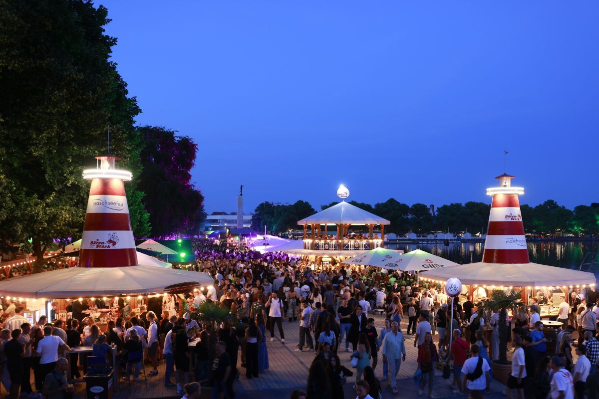 Crowd at an outdoor festival by a waterfront with tents, string lights, and two red-and-white lighthouse structures at dusk.