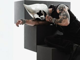 Person in a sleeveless black outfit leans over large black geometric blocks, wearing a shiny silver mask with cutouts and visible tattoos on the arm.