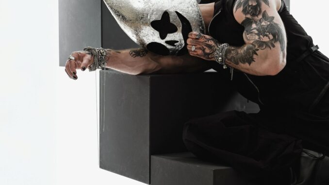 Person in a sleeveless black outfit leans over large black geometric blocks, wearing a shiny silver mask with cutouts and visible tattoos on the arm.