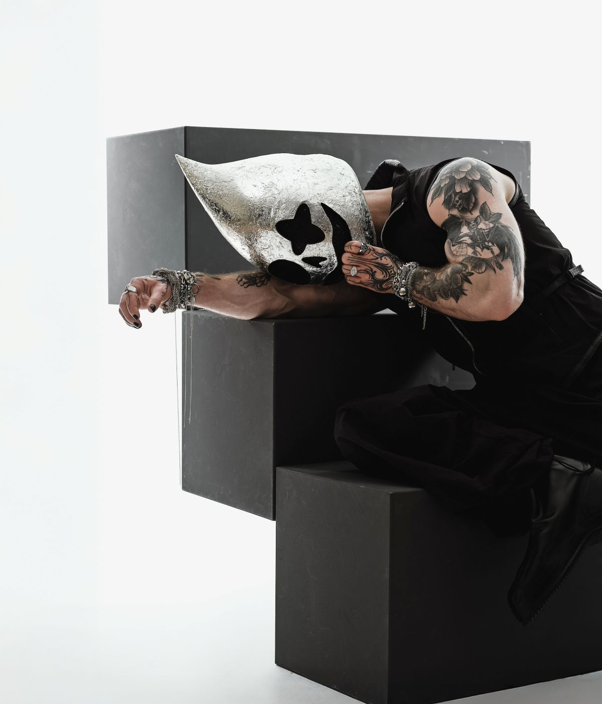 Person in a sleeveless black outfit leans over large black geometric blocks, wearing a shiny silver mask with cutouts and visible tattoos on the arm.