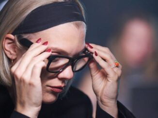 Woman with a blonde bob and black headband adjusts her glasses, focused downward.