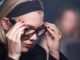 Woman with a blonde bob and black headband adjusts her glasses, focused downward.