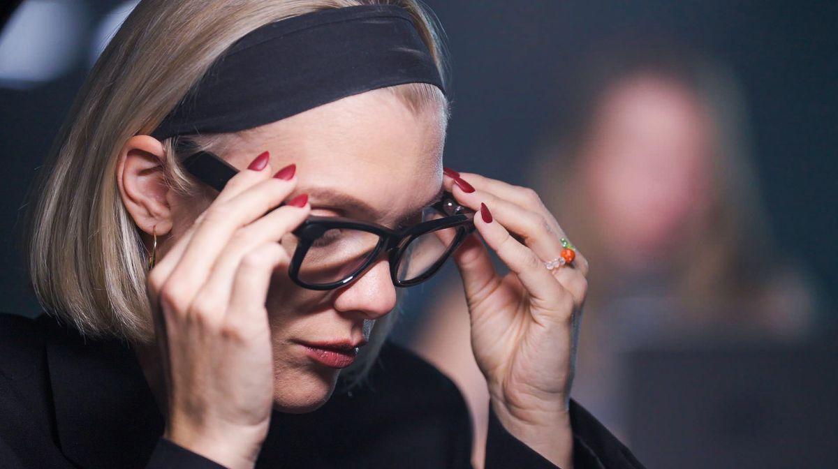 Woman with a blonde bob and black headband adjusts her glasses, focused downward.