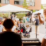 Smiling woman in a white dress sings into a microphone at an outdoor live event; pianist visible in the foreground while a crowd and white tents bustle in the background.