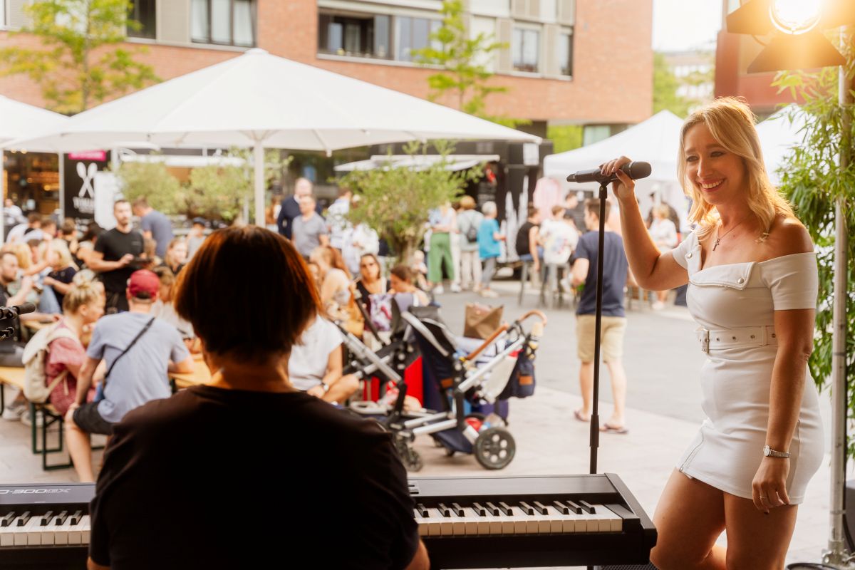 Smiling woman in a white dress sings into a microphone at an outdoor live event; pianist visible in the foreground while a crowd and white tents bustle in the background.
