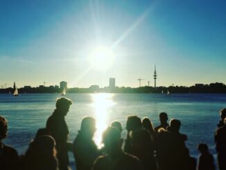 Silhouetted crowd by a sunlit lake watching a bright sunset with boats and a distant city skyline.