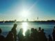 Silhouetted crowd by a sunlit lake watching a bright sunset with boats and a distant city skyline.