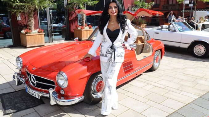 Woman in a white suit with black top posing beside a red vintage Mercedes convertible at a sunny car show street scene.