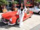 Woman in a white suit with black top posing beside a red vintage Mercedes convertible at a sunny car show street scene.