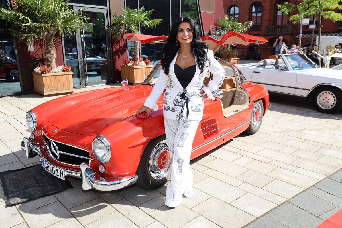 Woman in a white suit with black top posing beside a red vintage Mercedes convertible at a sunny car show street scene.