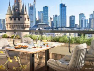 Rooftop dining table with cushioned chairs and plates of food, overlooking a modern city skyline with a castle-like turret in the foreground.