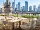 Rooftop dining table with cushioned chairs and plates of food, overlooking a modern city skyline with a castle-like turret in the foreground.