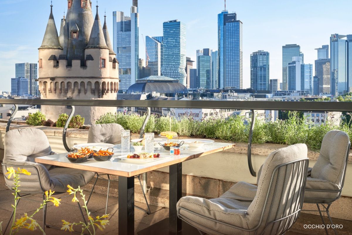 Rooftop dining table with cushioned chairs and plates of food, overlooking a modern city skyline with a castle-like turret in the foreground.